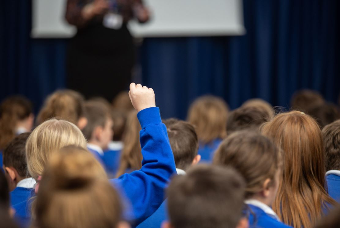 A child with their hand up and a school assembly 