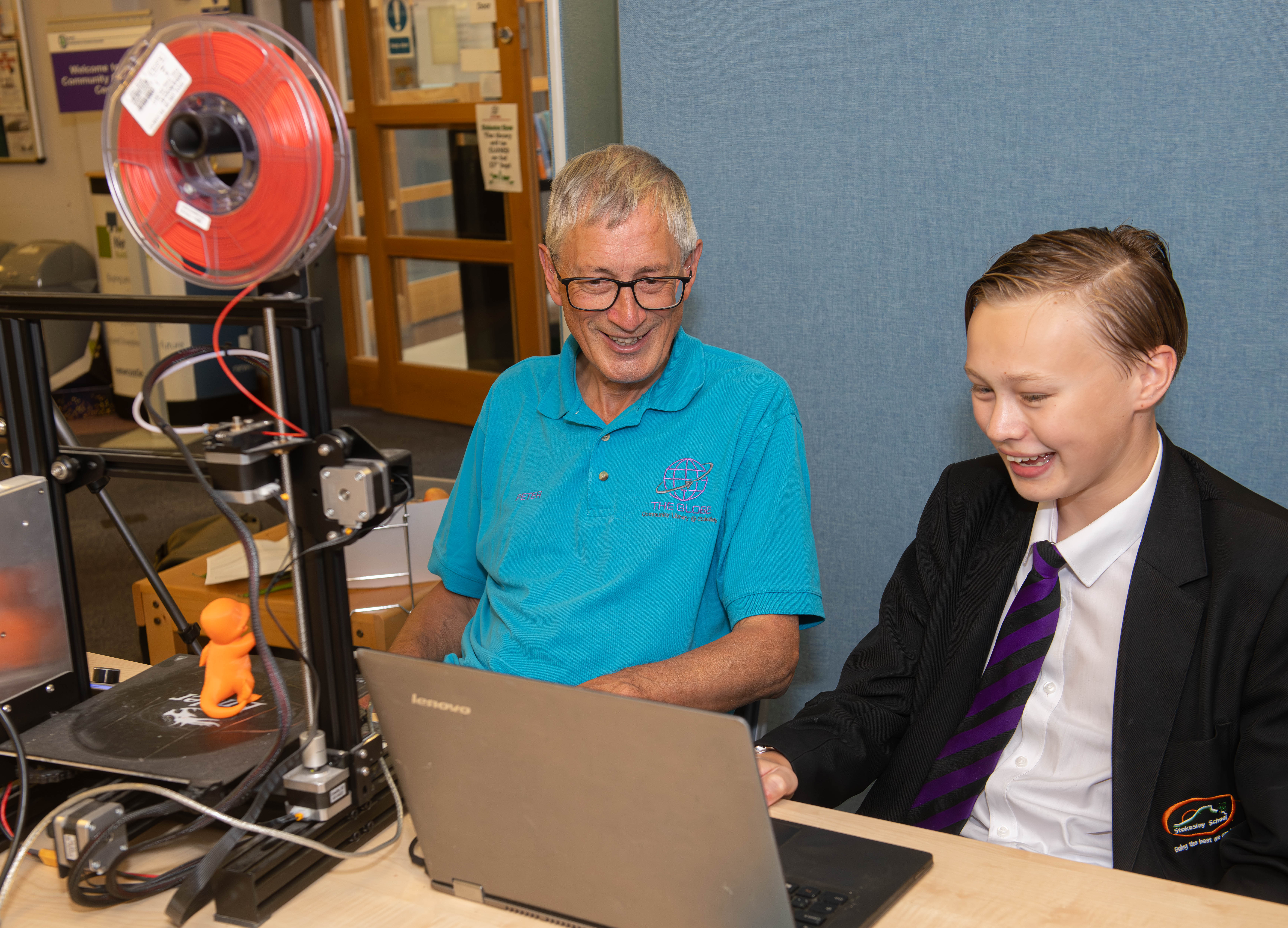 An IT buddy volunteer with a young child at a 3D printer and laptop