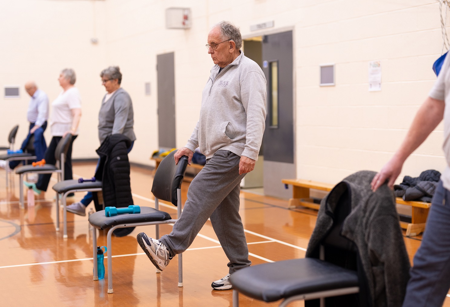A line of people balancing on one foot while holding the back of a chair as part of a session at Jennyfield Styan Community Wellbeing Hub