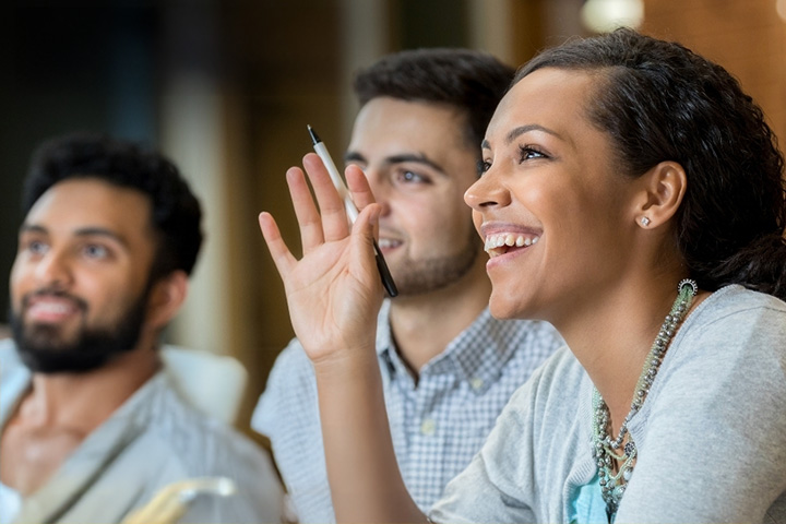Three people in a classroom, one with her hand raised.