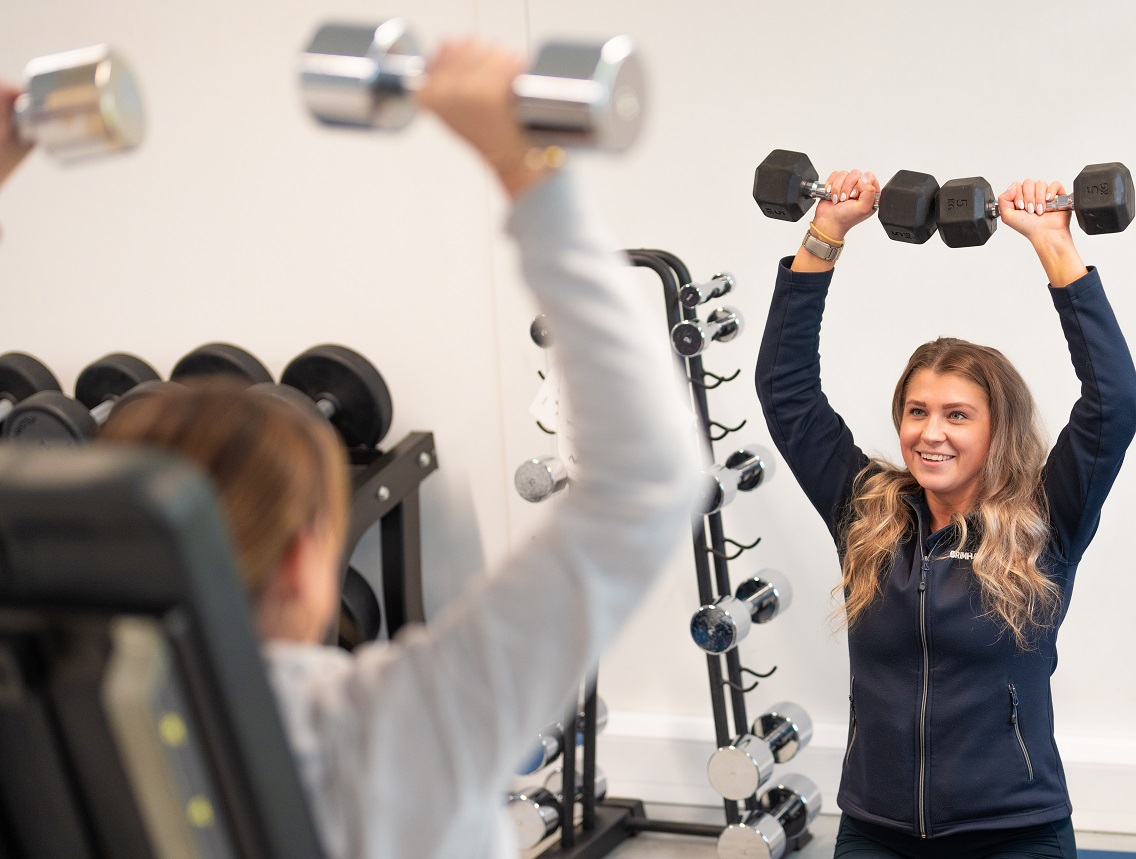 Two women lift weights, one facing the camera one with her back to it, in front of a room full of gym equipment.