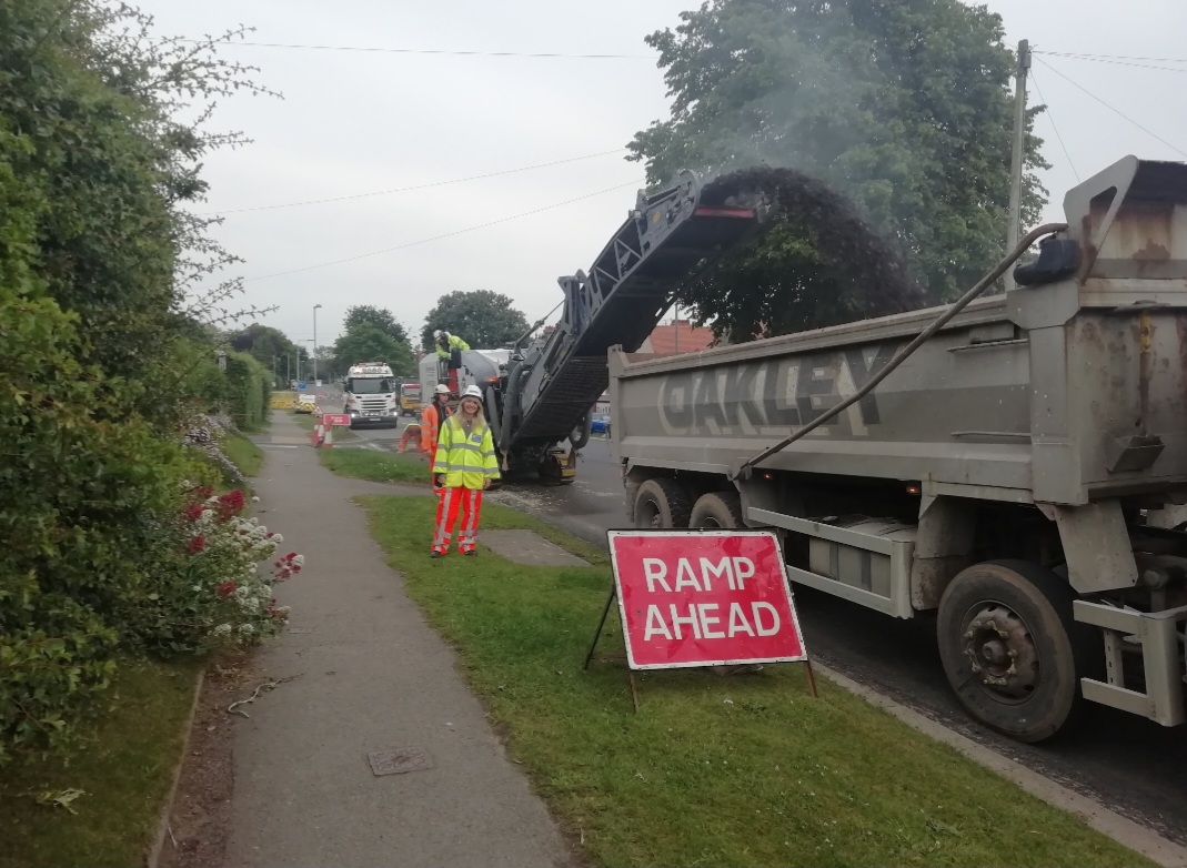 Adult in hi-viz standing on the pavement next to roadwork machinery and ramp ahead sign