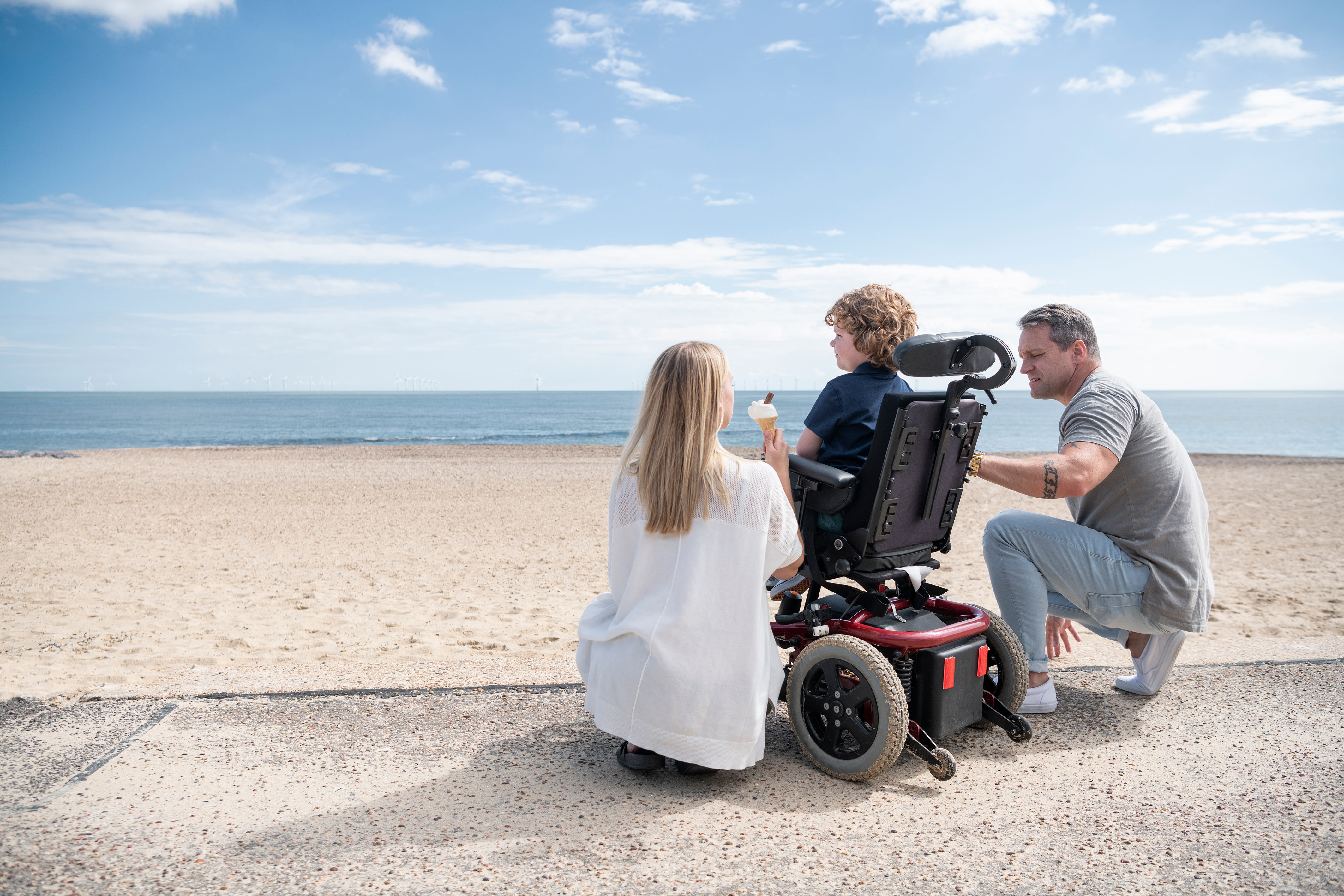 Two adults, one holding an ice cream with a child in a wheelchair in the middle at the seaside