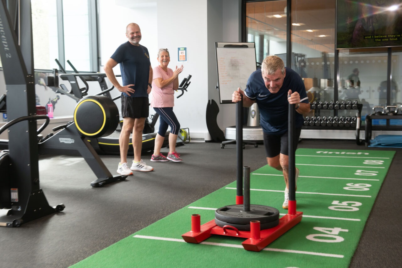 A man pushes a weighted resistance trolley in a gym while two other cheer him on.