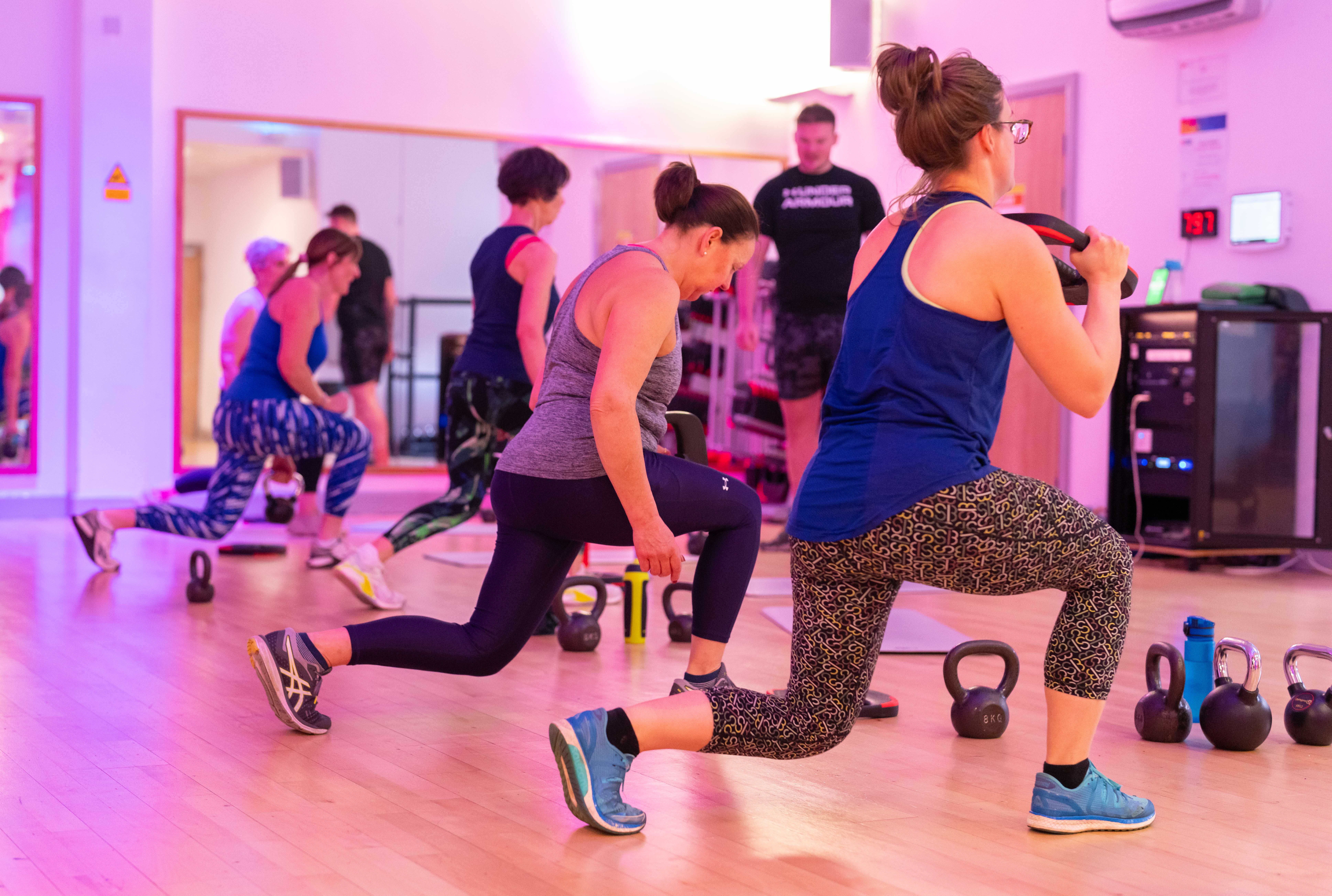 A group of women taking part in a kettlebells class while an instructor encourages them.