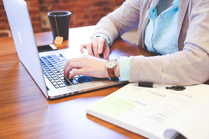 Person typing on a laptop with textbook and pen next to them.