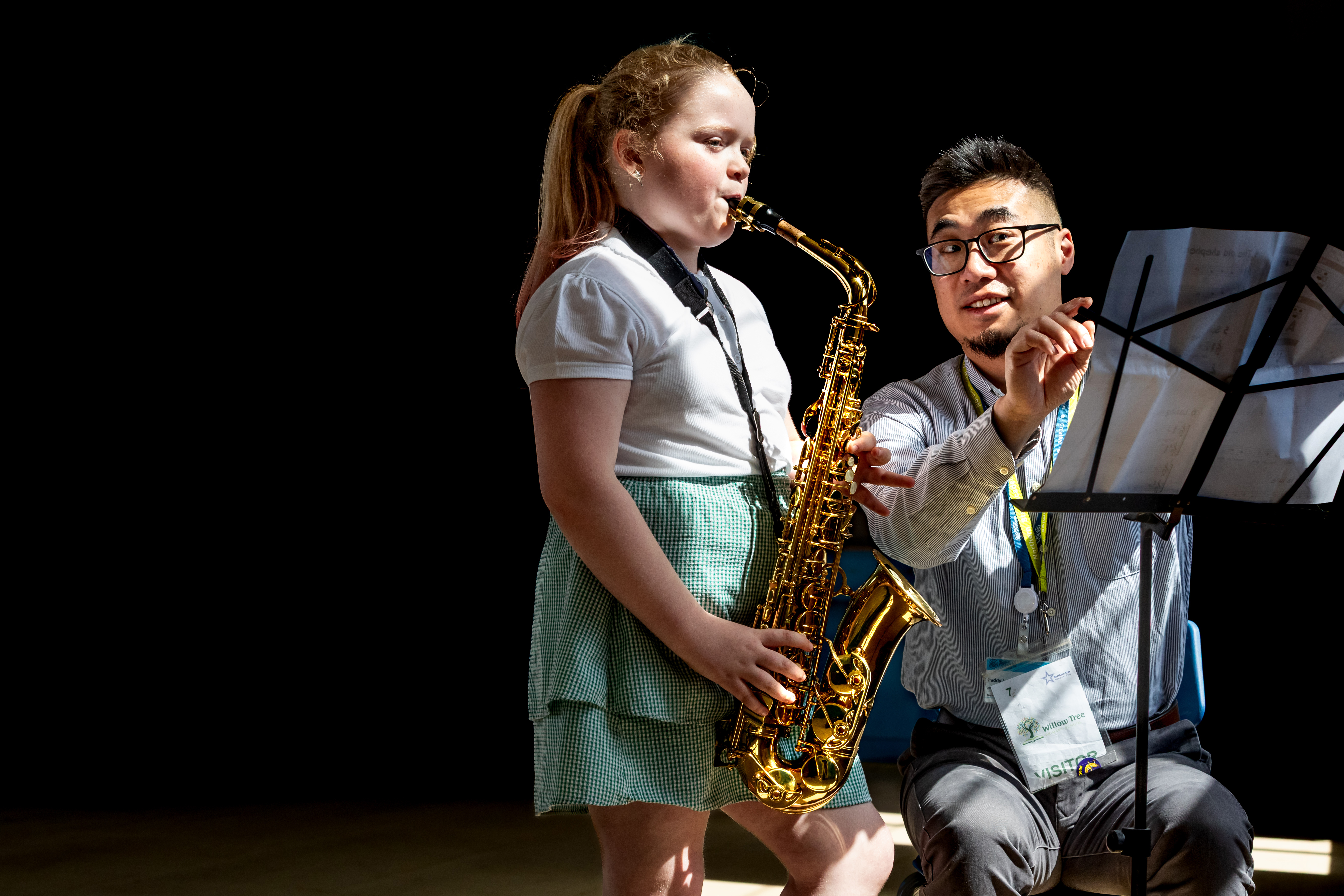 Child standing playing a saxophone with a teacher sitting beside her