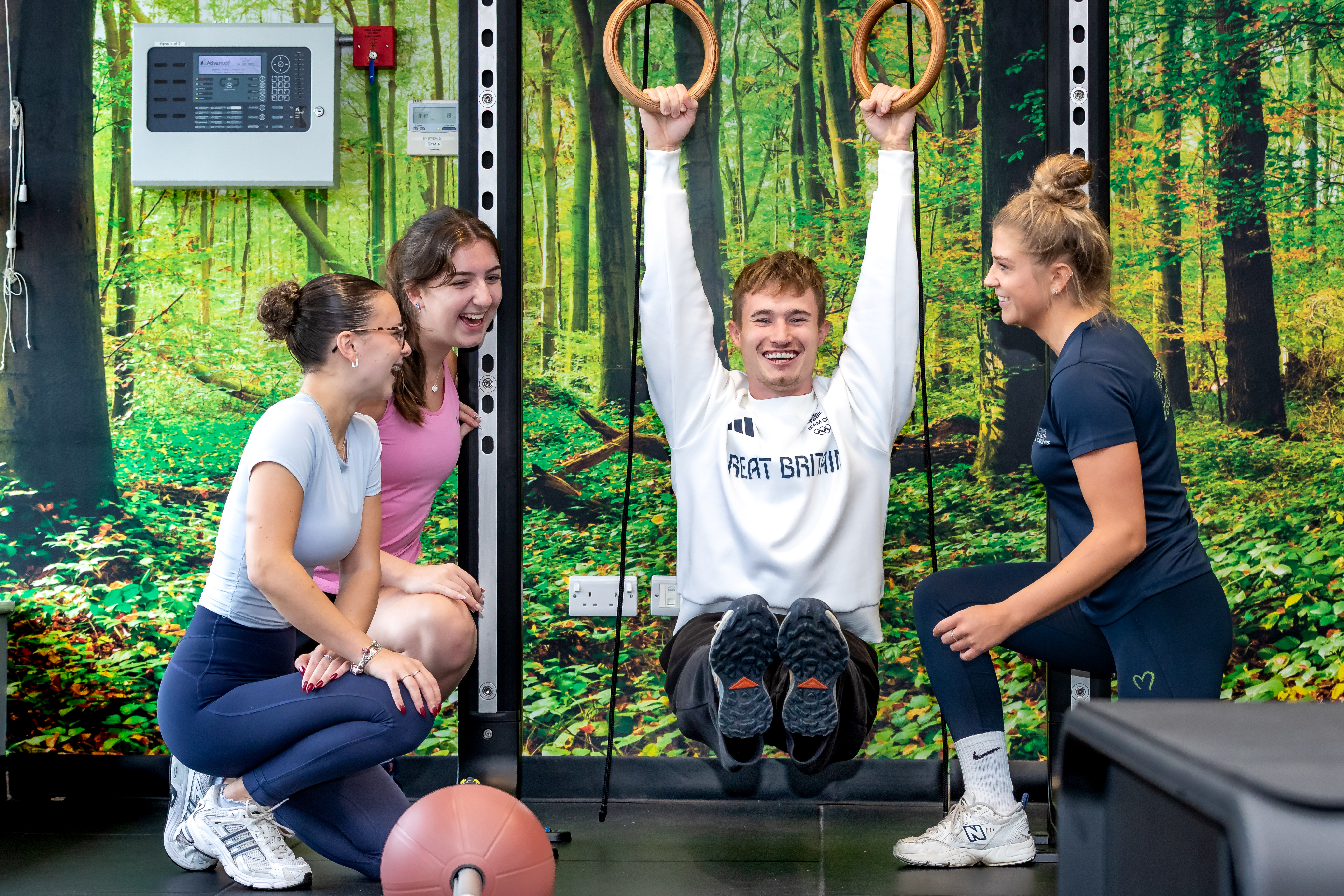 Four people exercising in a gym with a forest mural with Jack Laugher MBE using gymnastic rings.