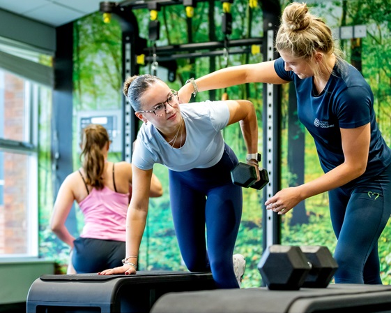 An instructor at Ripon Leisure and Wellbeing Hub - The Jack Laugher Centre helps a woman with her form while lifting weights. Another woman works out in the background.