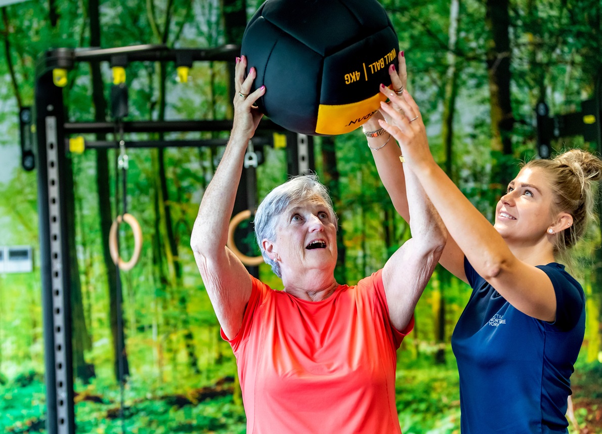 A lady lifts a 4kg wall ball above her head while an instructor helps to hold it steady.