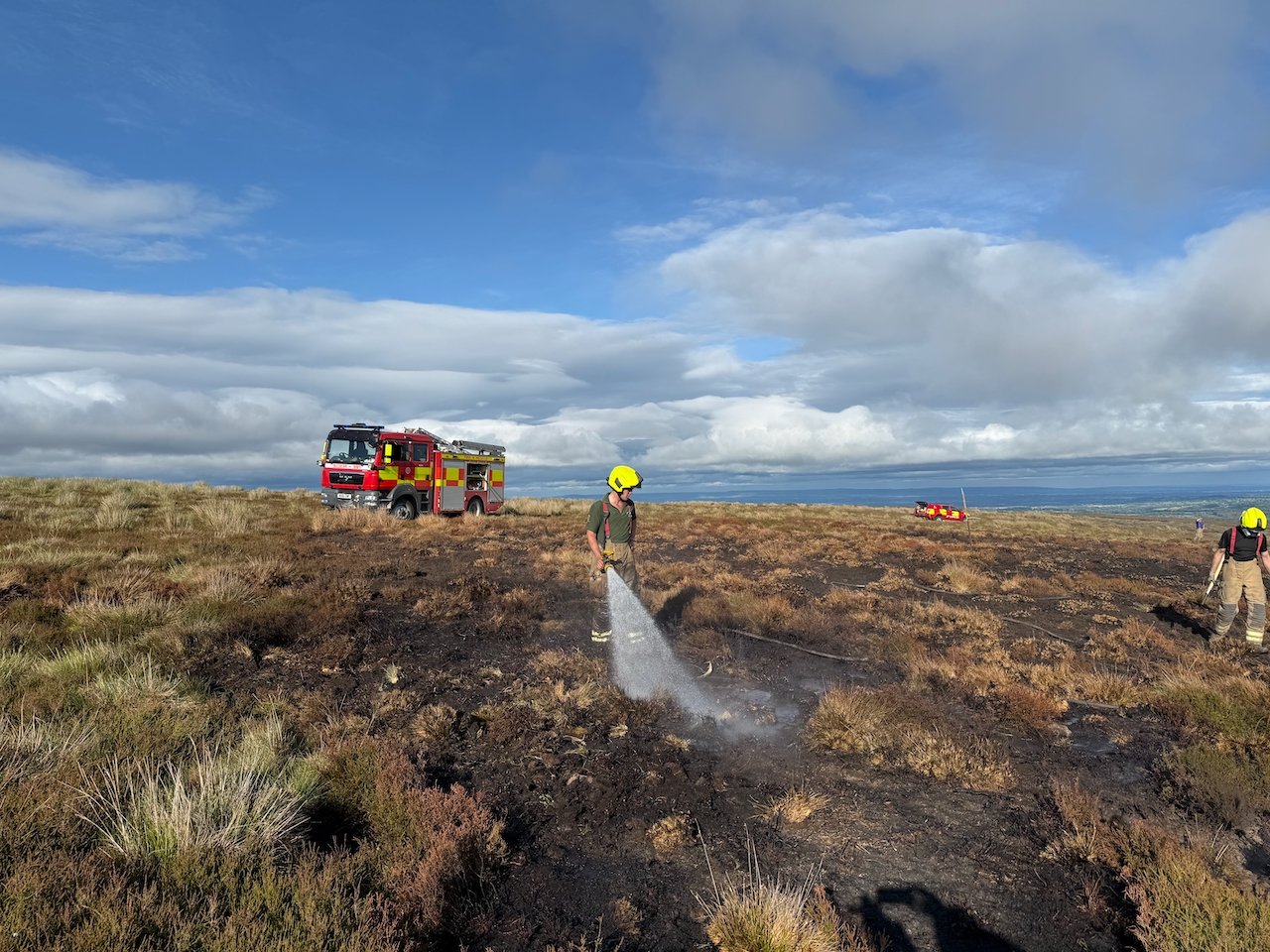 A fireman soaks the burnt ground on top of the moor using a firehouse. A fire engine can be seen in the background.