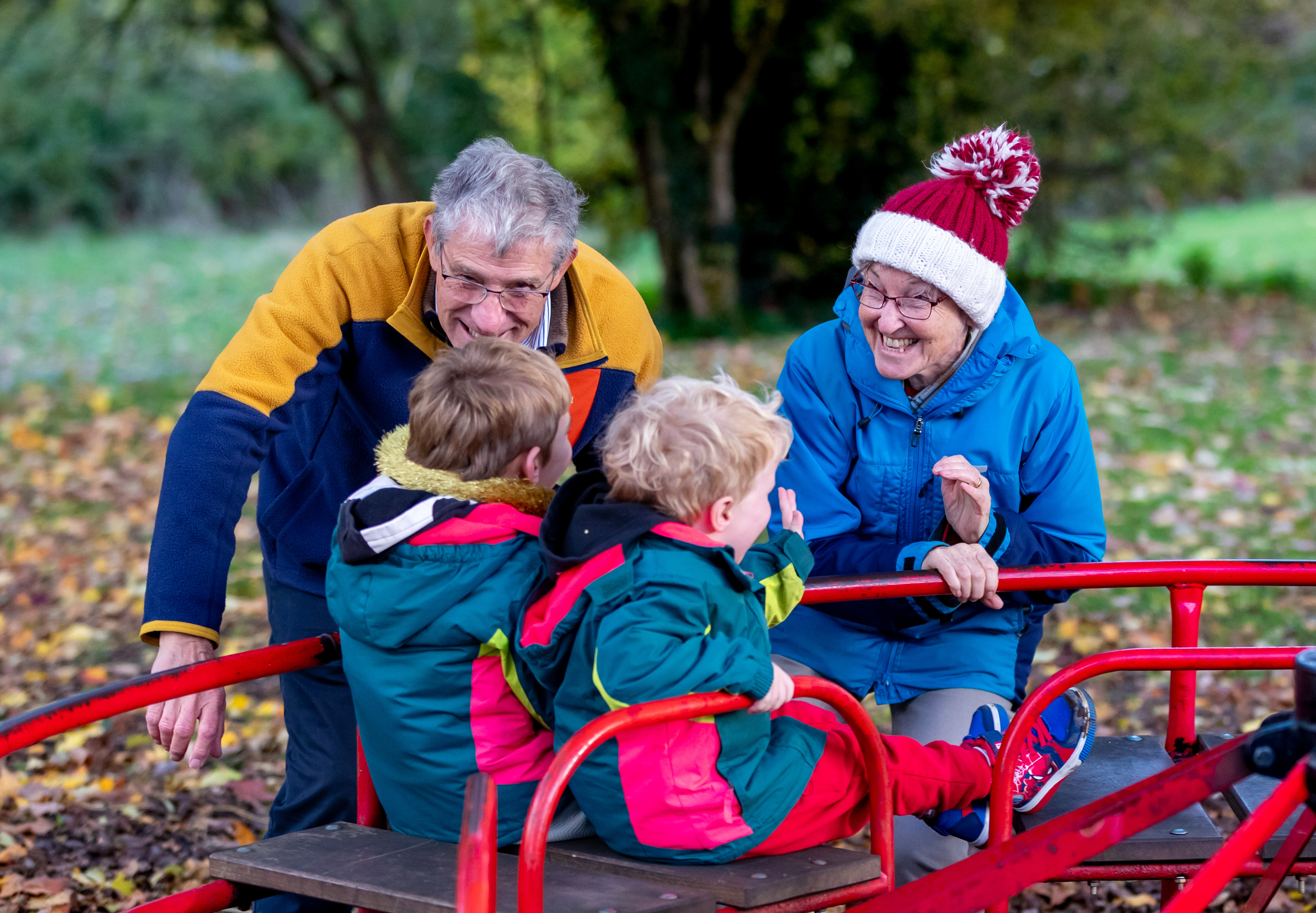 Two adults with two young children on a red roundabout outside on an autumn day