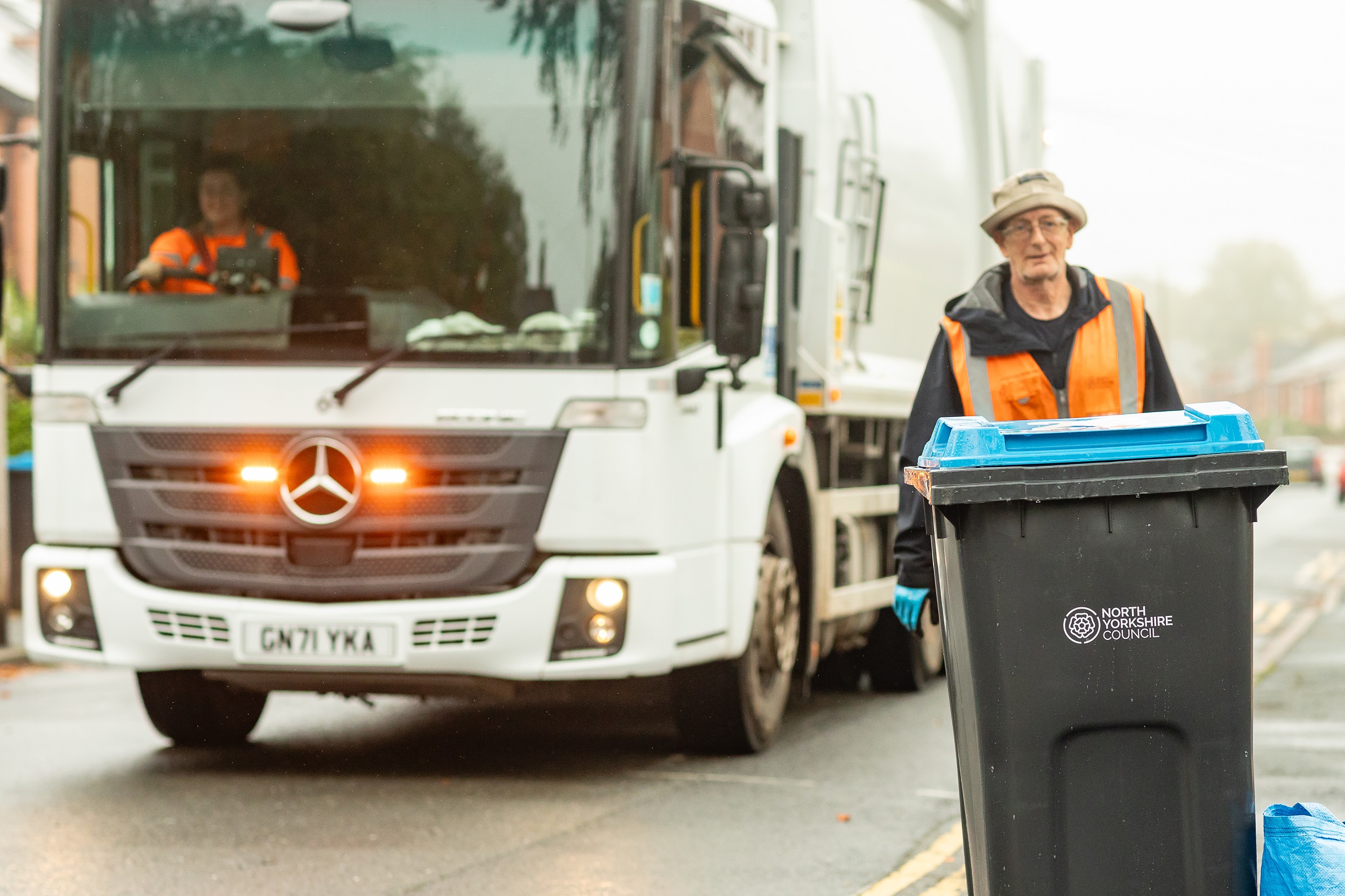 Recycling bin being collected with a recycling vehicle behind.