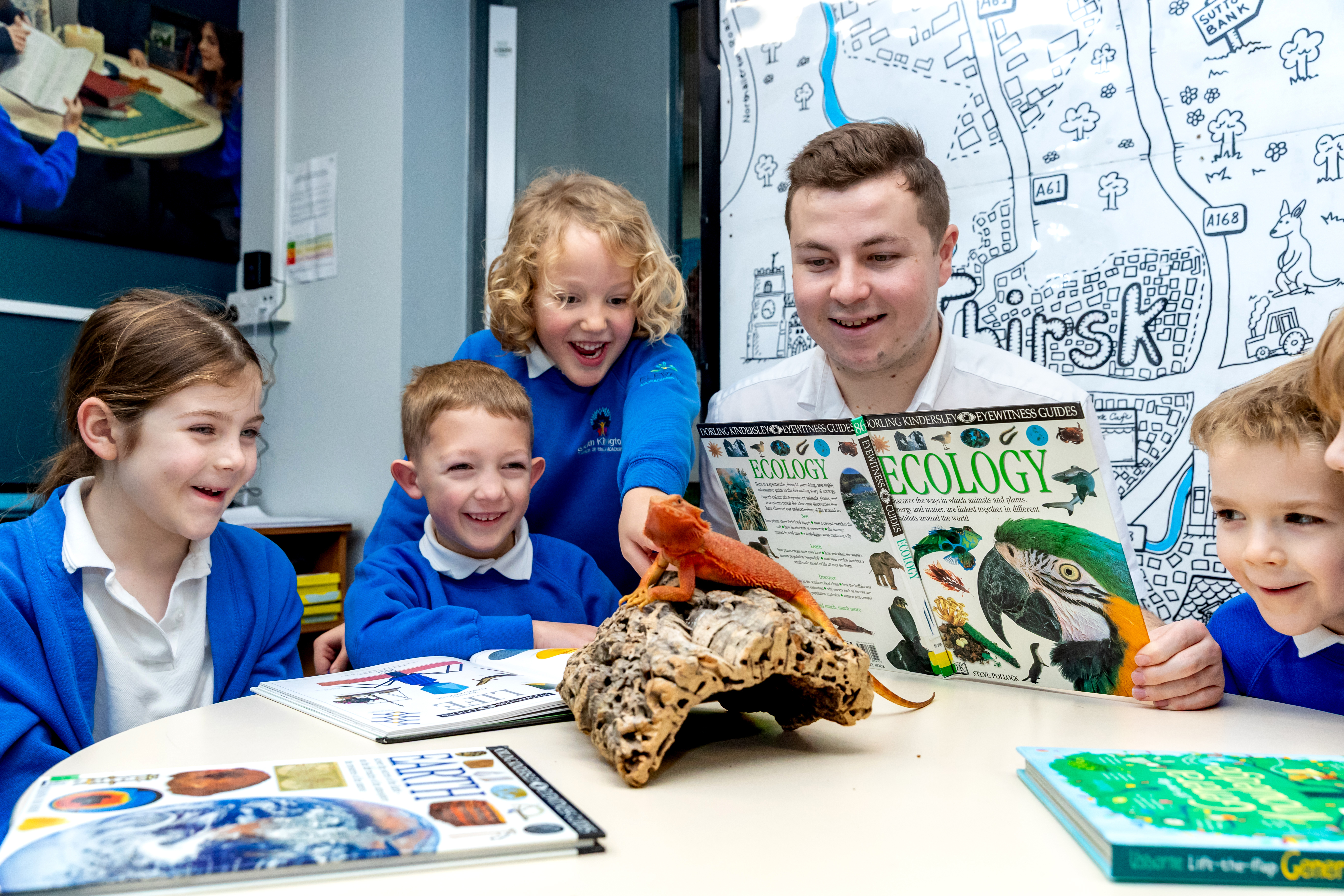 Ben and several children in their school uniform sitting at a table reading a book on Ecology with a reptile sitting on a piece of wood in front of them