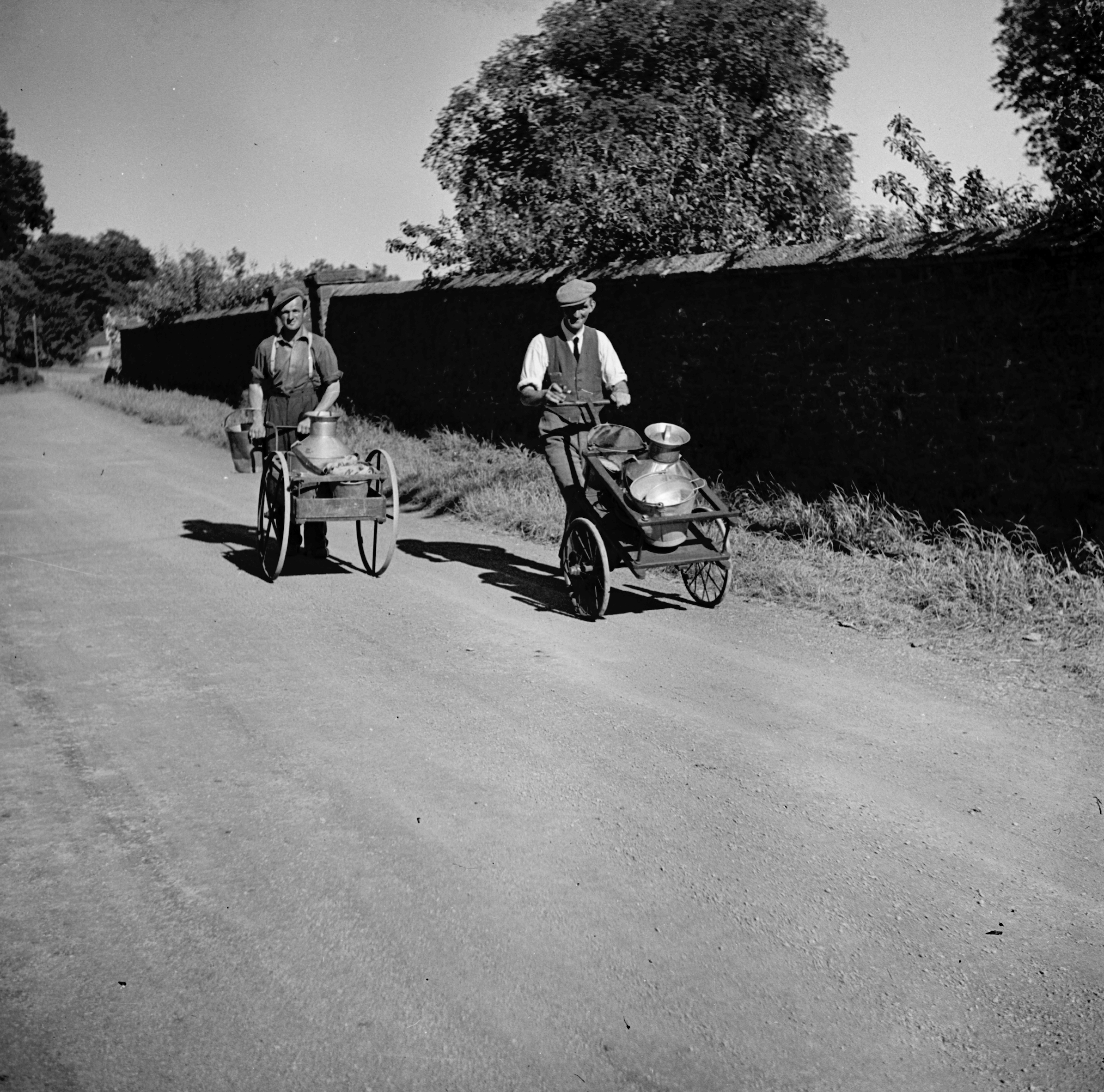Black and white photo of two adults collecting milk in Hawes. They use hand carts to transport the budget cans along the road (mid-20th-century). From the Bertram Unné photographic collection.