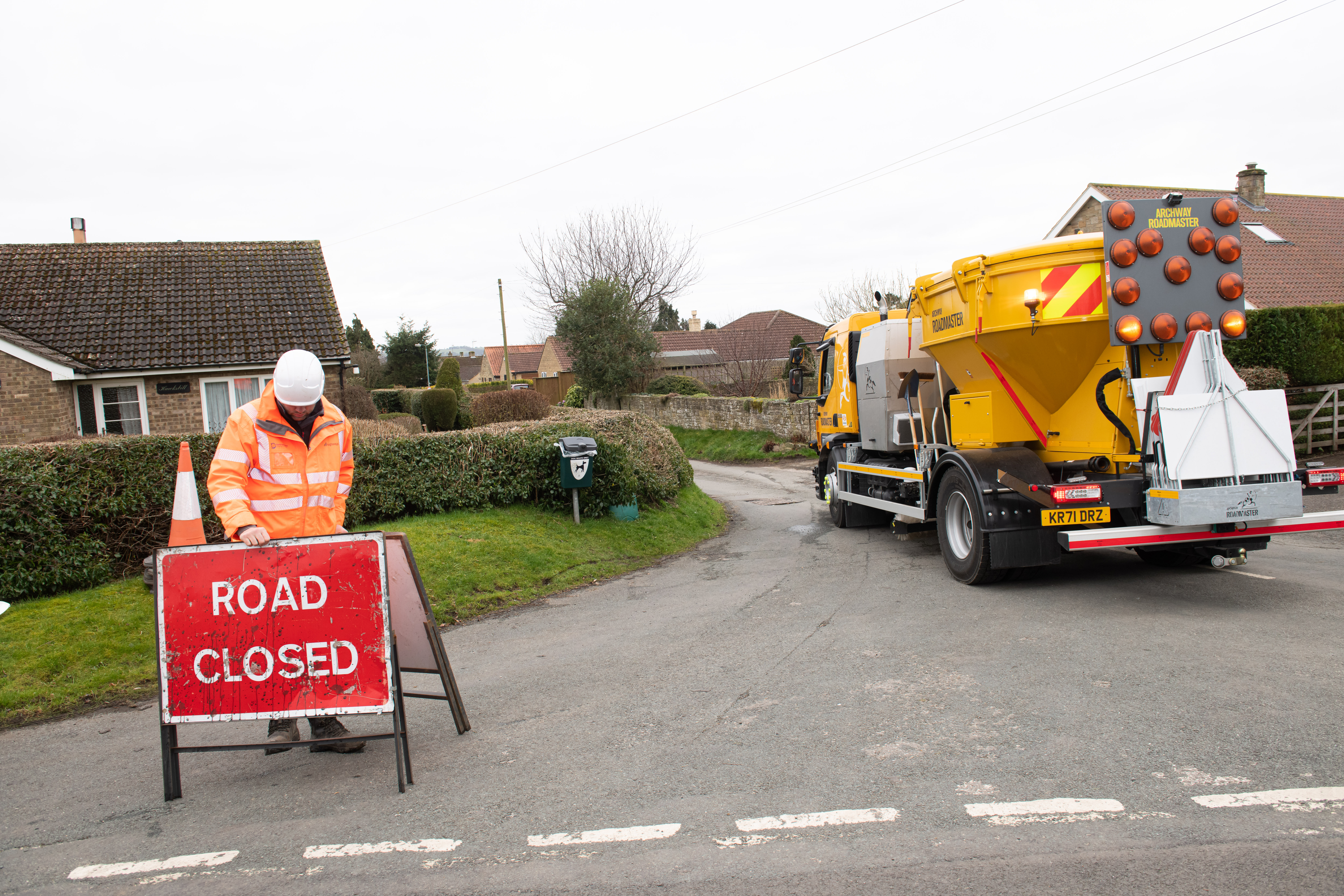 Person in hi-viz placing a road closed sign at the junction of a road in front of a highways vehicle