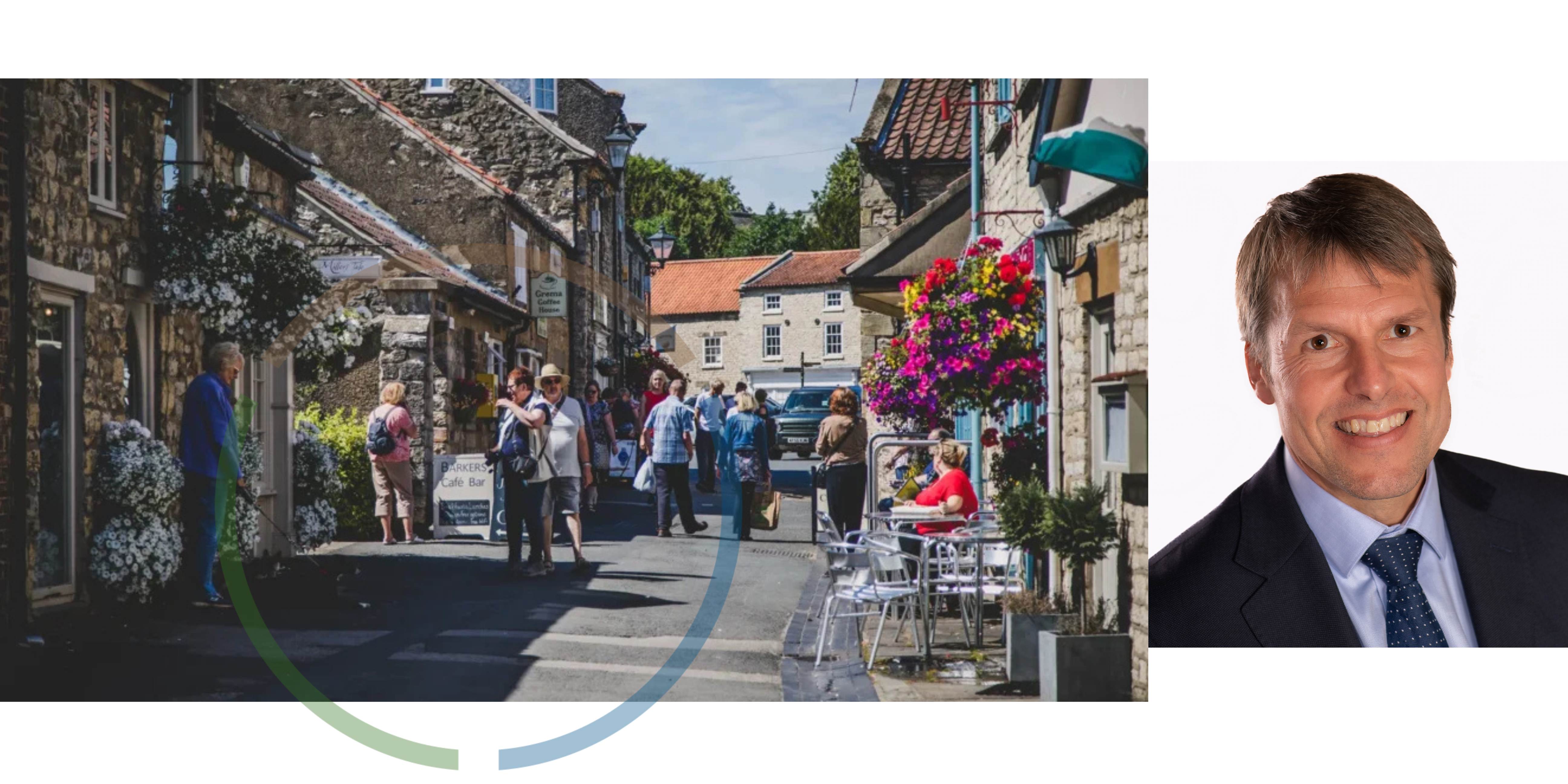 Stone brick shops with colourful hanging baskets and several groups of people walking down the middle and a photo of Nic Harne