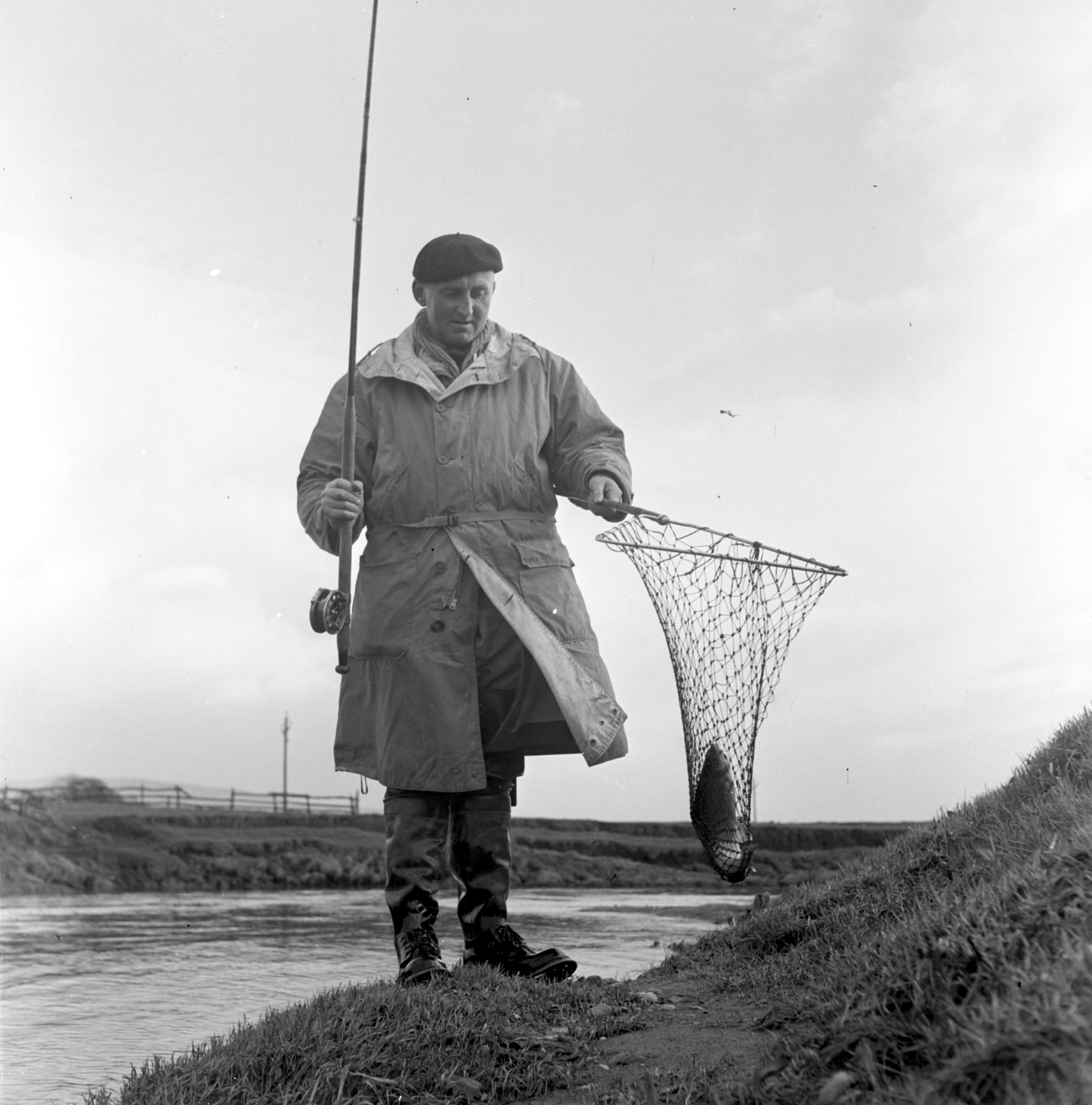 Black and white photo of a fisherman assessing his catch on the banks of the River Aire in the 20th century. From the Bertram Unné photographic collection.