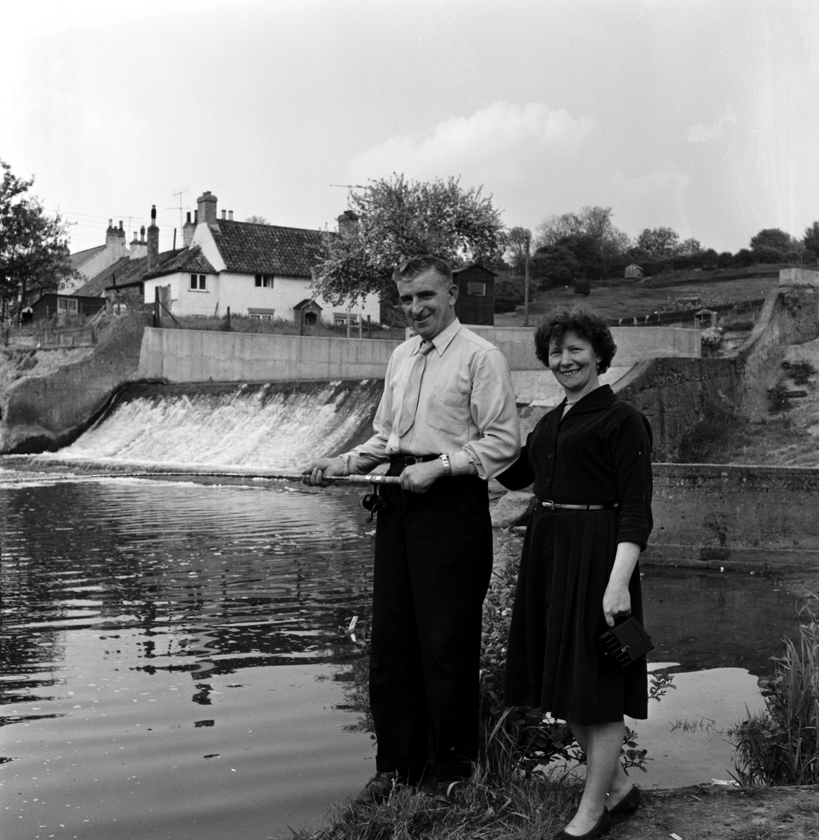 Black and white photo of a couple pictured fishing together on the River Leven during the 20th century. From the Bertram Unné photographic collection.