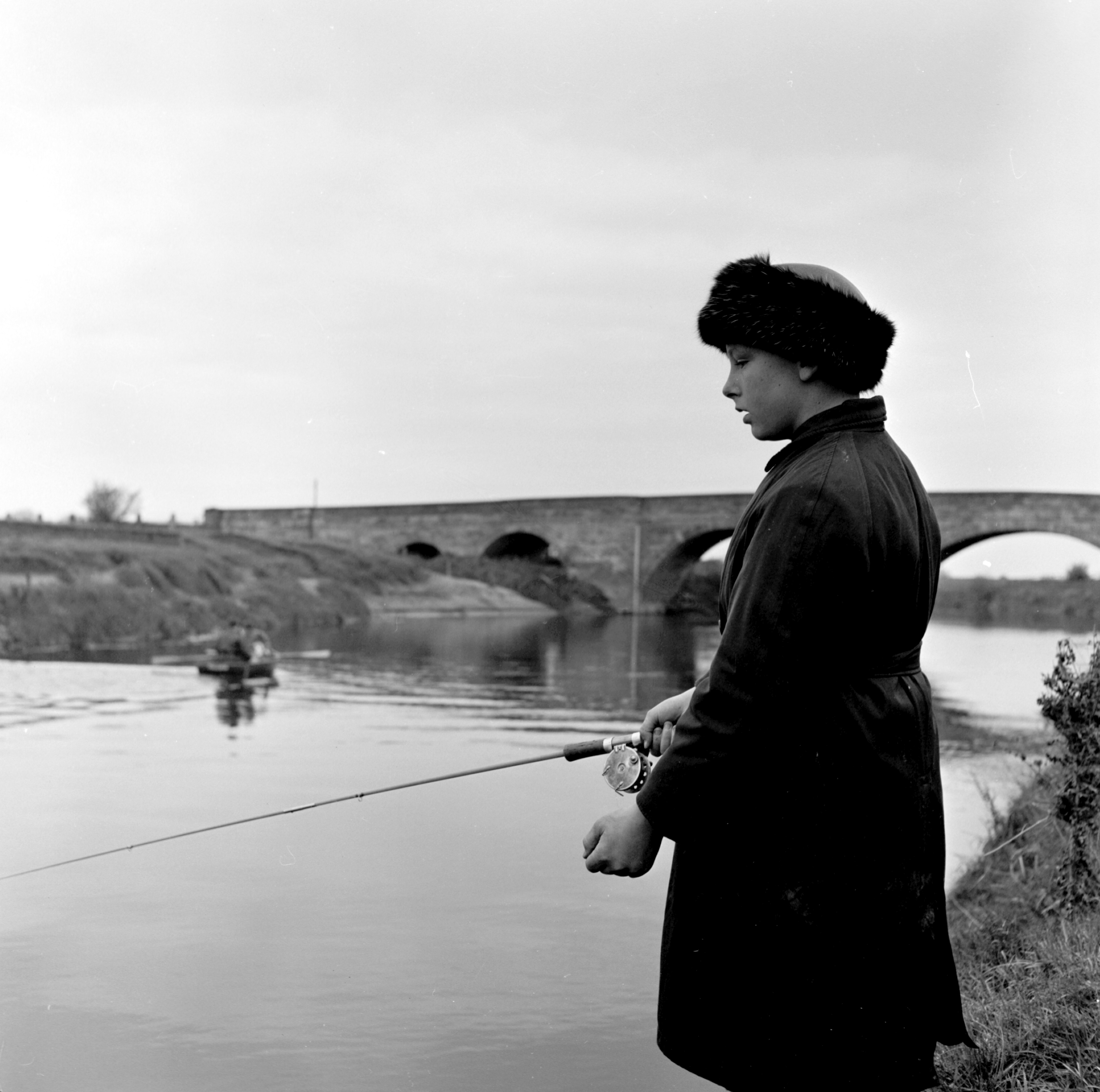 Black and white photo of a woman fishing along from Skipton-on-Swale bridge in the 20th century. From the Bertram Unné photographic collection.