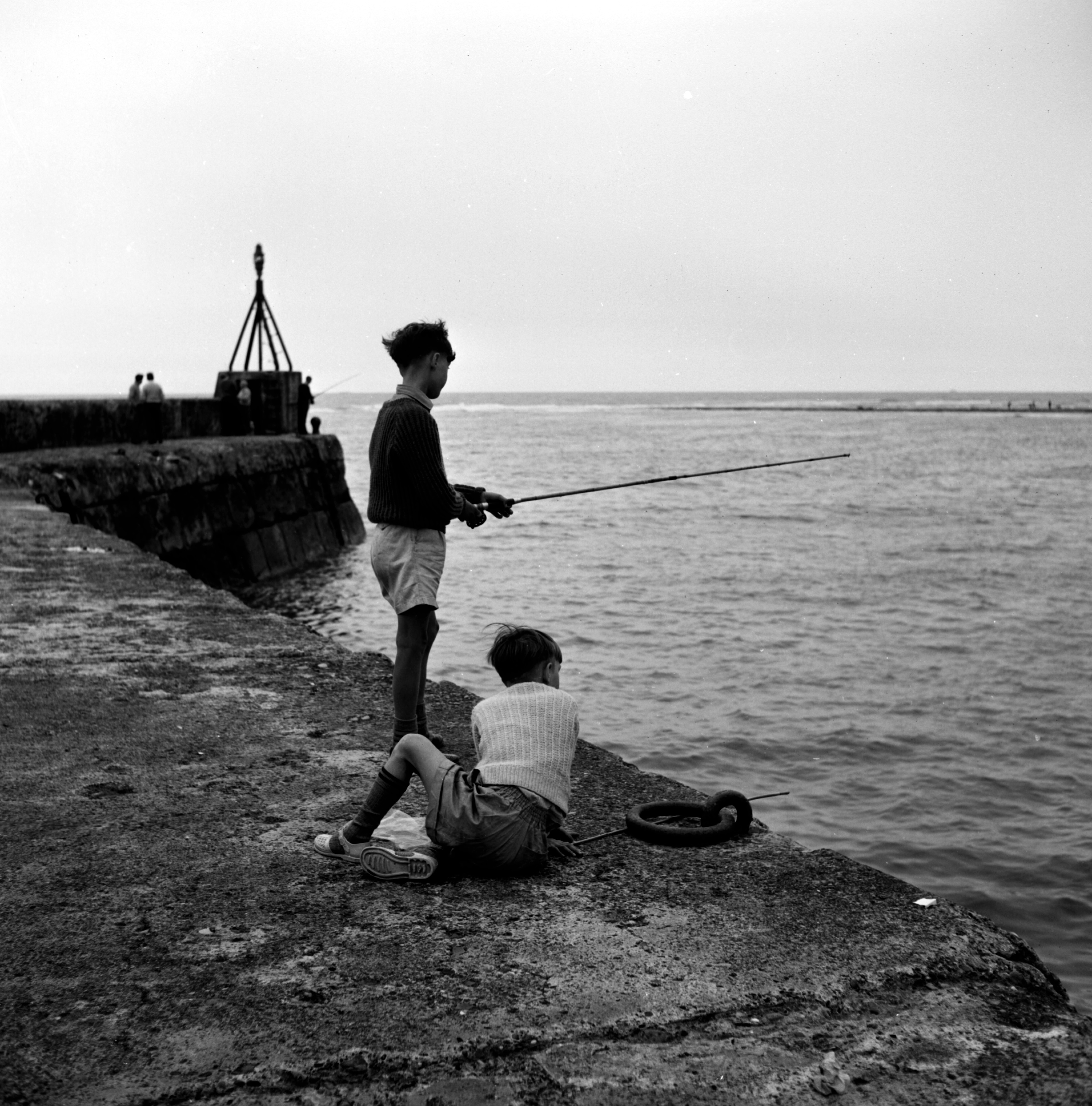 Black and white photo of youngsters trying their hand at sea fishing off Whitby Pier in the 20th century. From the Bertram Unné photographic collection.