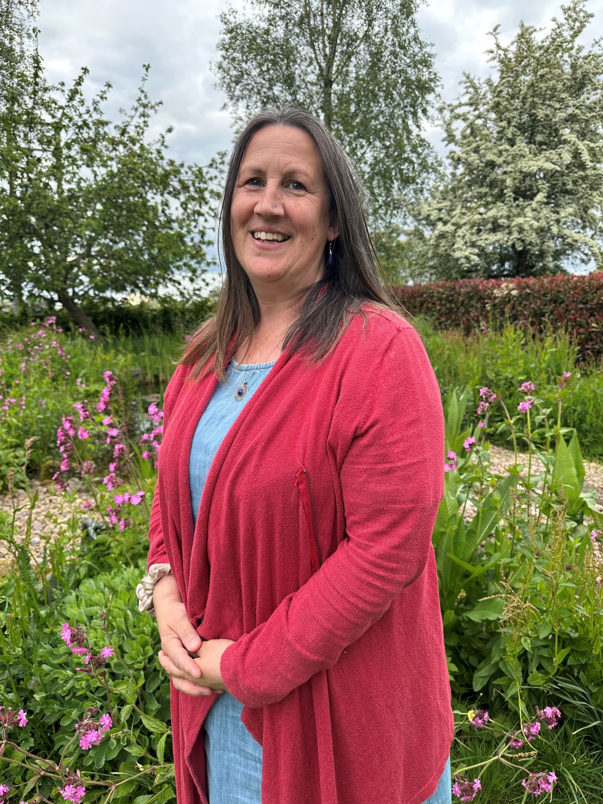 A photo of Helen Goodwin standing outside in front of flowers, hedging and trees