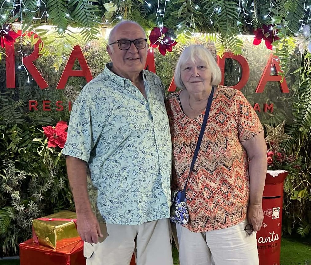 Photo of Denise and Colin standing in front of wrapped presents and under fairy lights