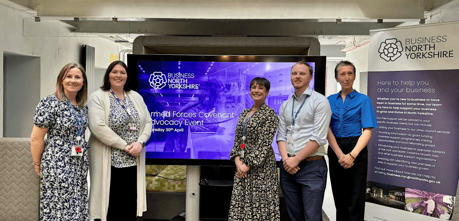 A group of people standing smiling in front of a large screen displaying the details of the Armed Forces Covenant event