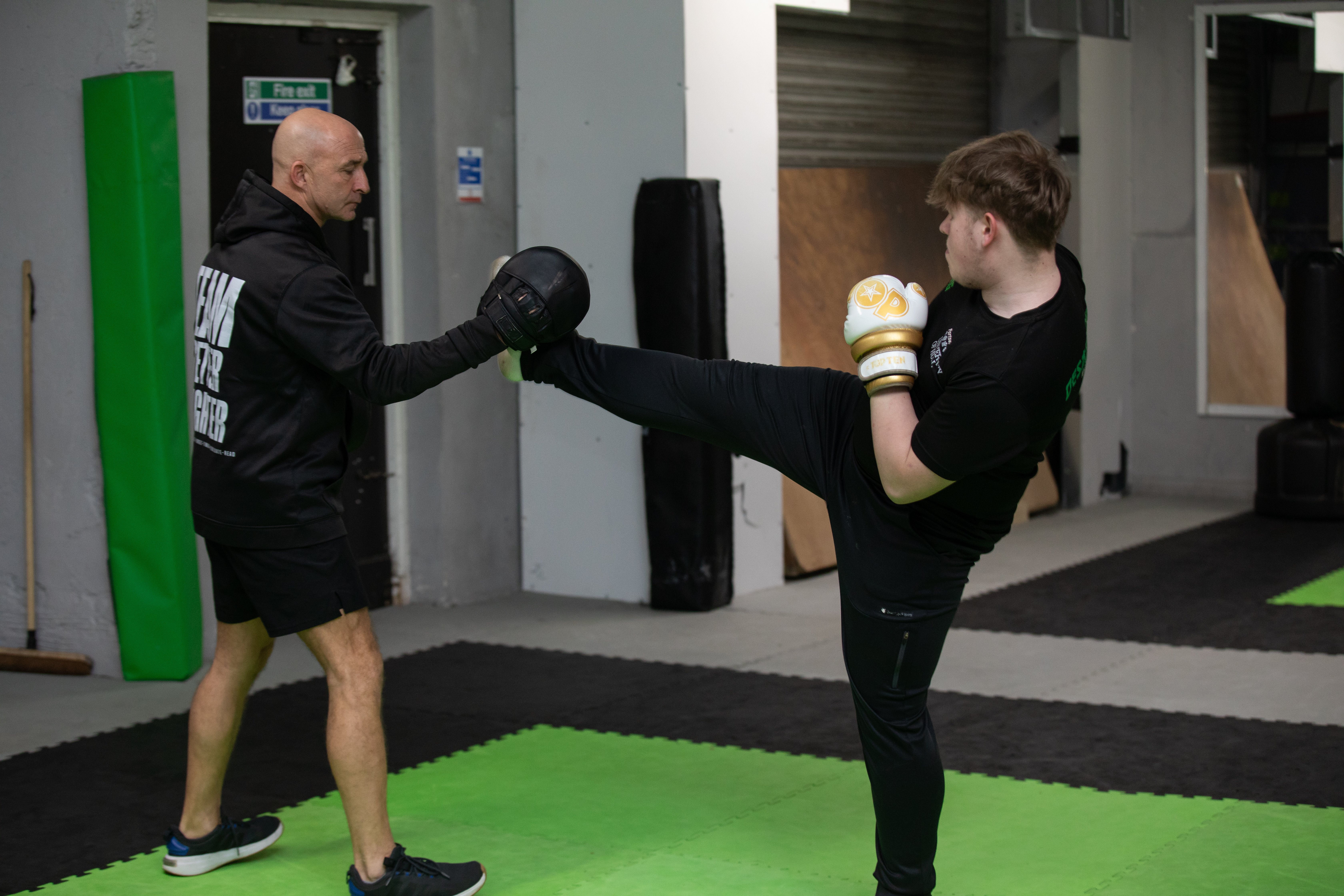 An adult doing a high martial arts kick reaching the pad on the hand of another adult