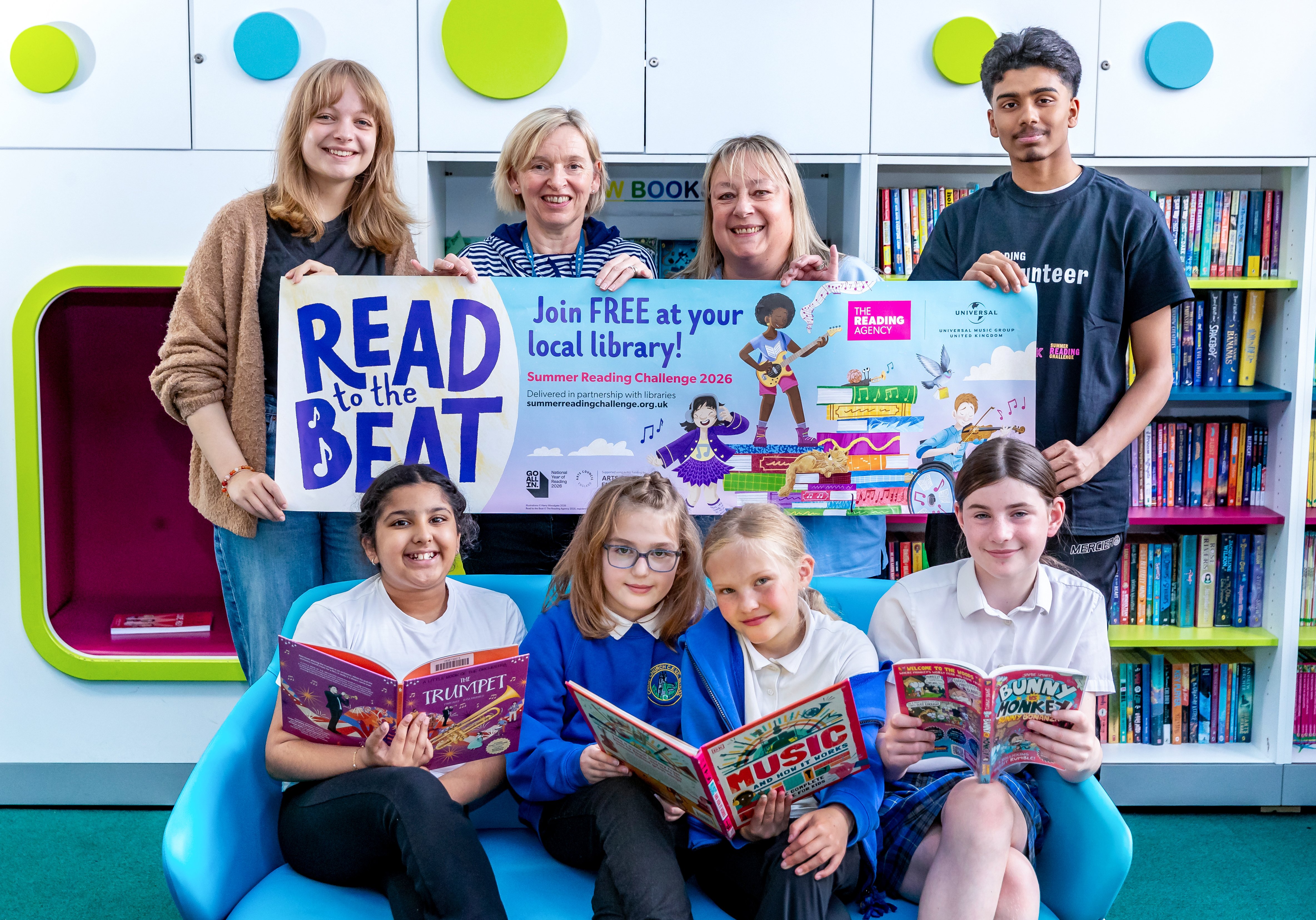 Small group of school children sitting on chairs holding books and some adults standing behind holding a read to the beat banner