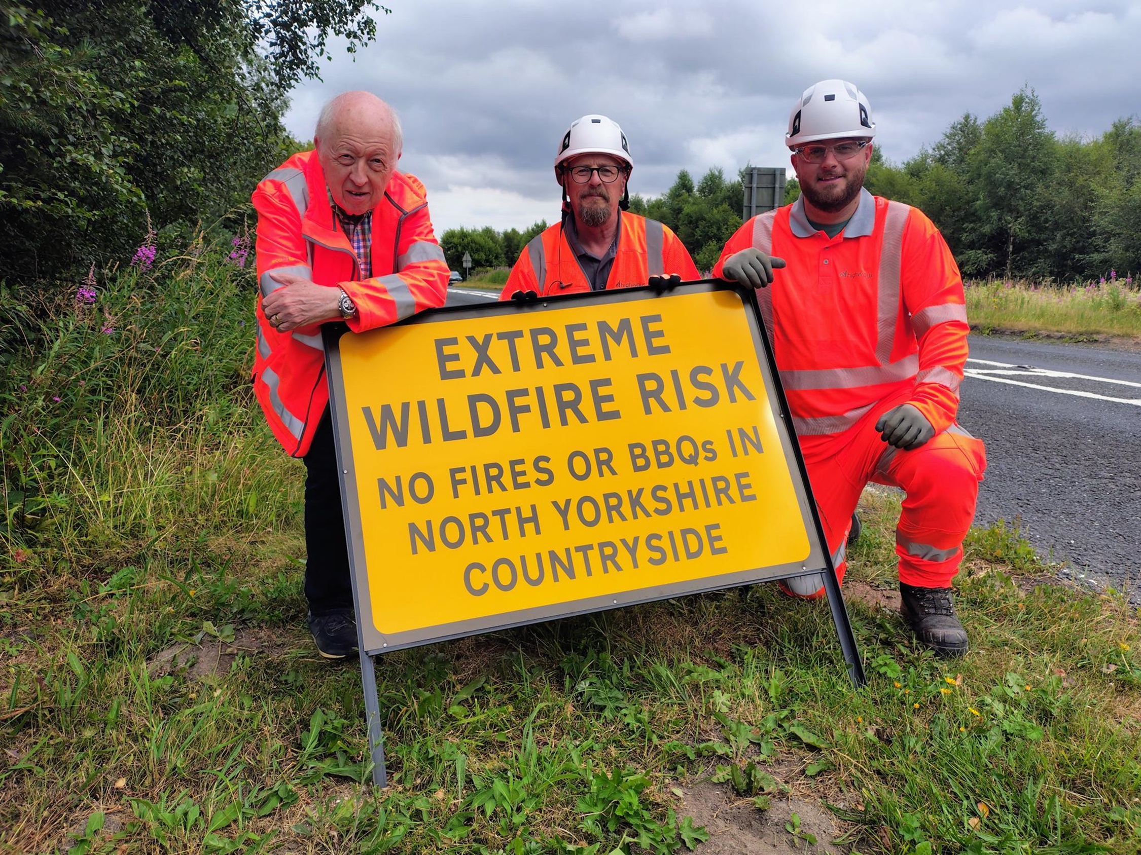 Three adults wearing hi viz clothing behind a wildlife warning sign on a grass verge