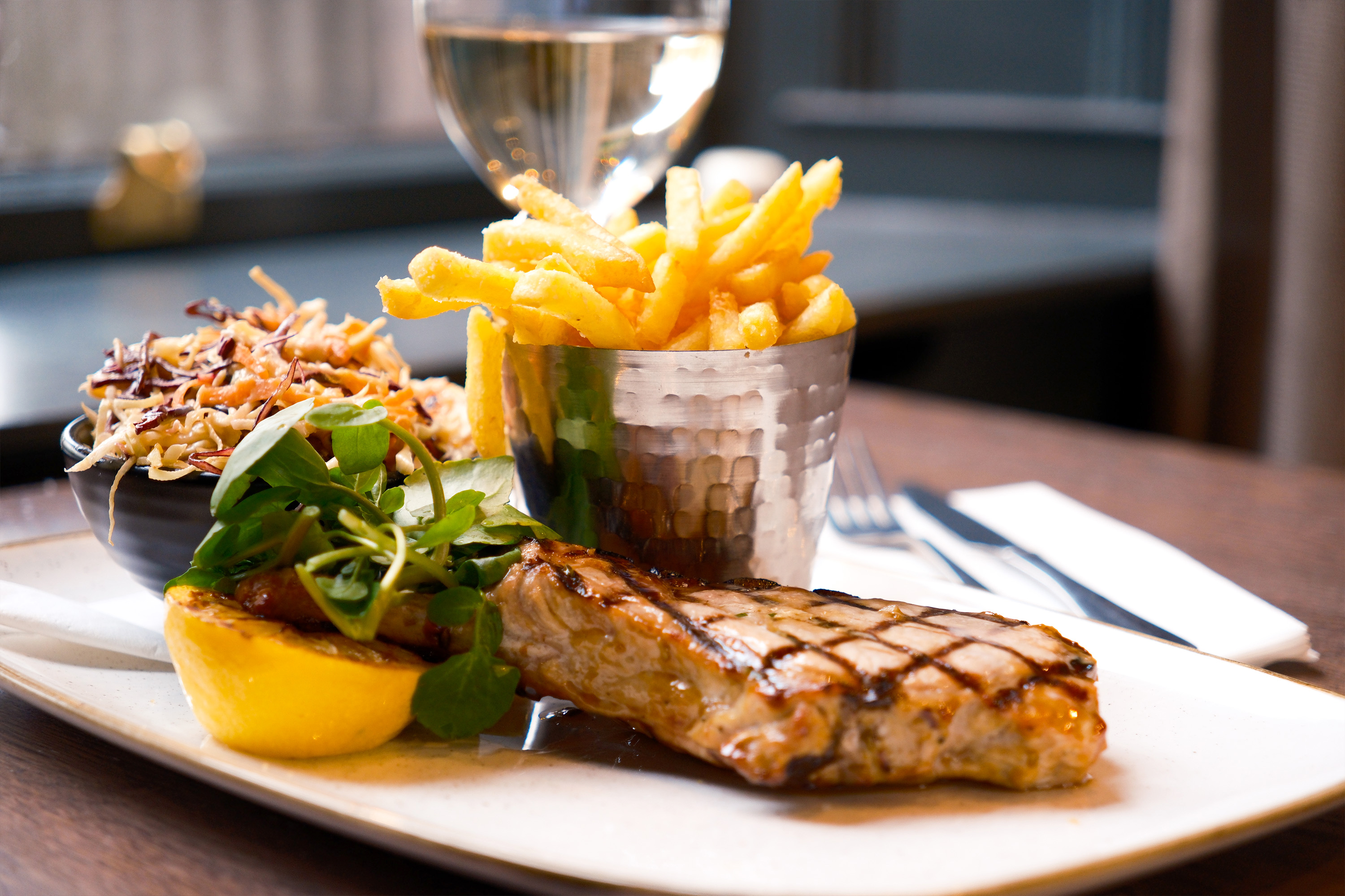 White dinner plate with meat, a silver container with skinny chips, a pot of coleslaw and a lemon and garnish