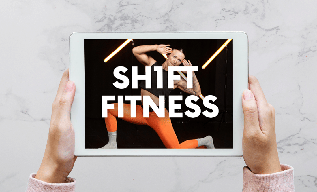 Hands holding a tablet which has a Shift Fitness logo on it over someone taking part in an exercise class.