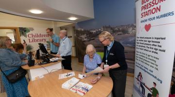 Image showing a person having their blood pressure taken happily chatting with a staff member whilst a group of people chat in the background.