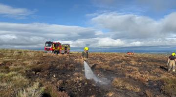 A fireman soaks the burnt ground on top of the moor using a firehouse. A fire engine can be seen in the background.