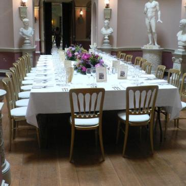 A long table with chairs and floral centre pieces and statues in the background. 