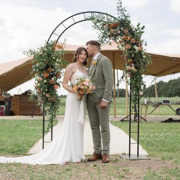 A wedding couple stood under a wire arch in a field
