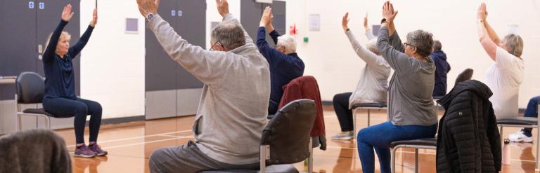 A group of people and an instructor sitting down with arms raised as part of a chair-based session at Jennyfield Styan Community Wellbeing Hub