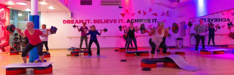 A group take part in an exercise class, squatting while carrying barbells.