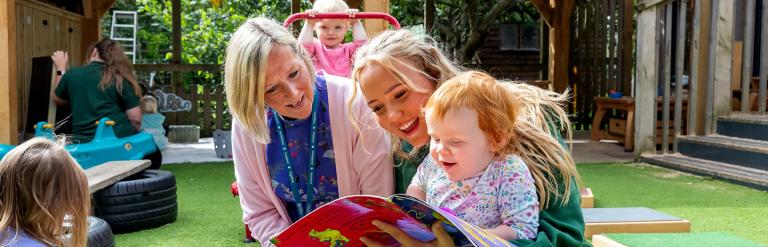 Couple of adults with nursery children sitting outside reading a book whilst a child is on a small trampoline behind