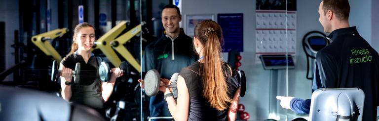 A lady working out in the gym with a personal trainer in front of the mirror.