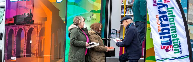 Three people outside the mobile library holding books.