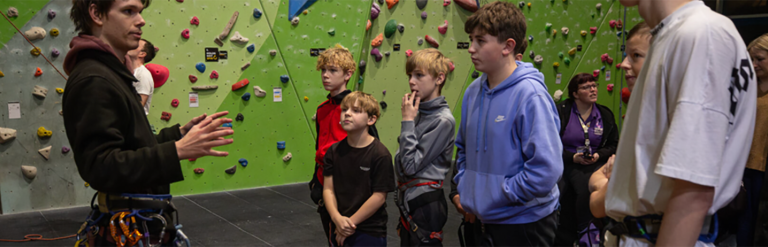 An adult and several children listening to the instructor beside an indoor climbing wall