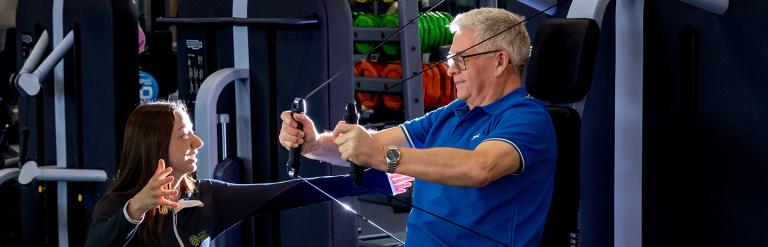 An adult on exercise equipment with an instructor giving instructions on how to use it