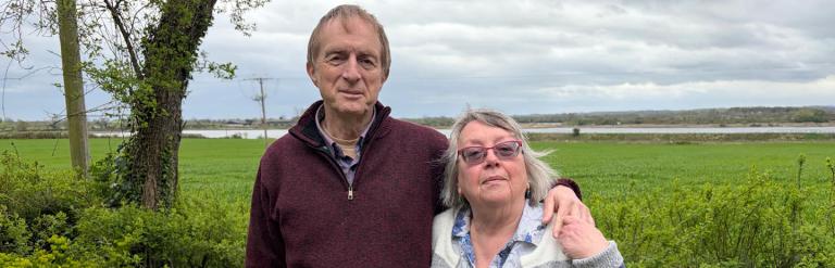 Couple standing together outside on a cloudy day with a greenery and water behind them