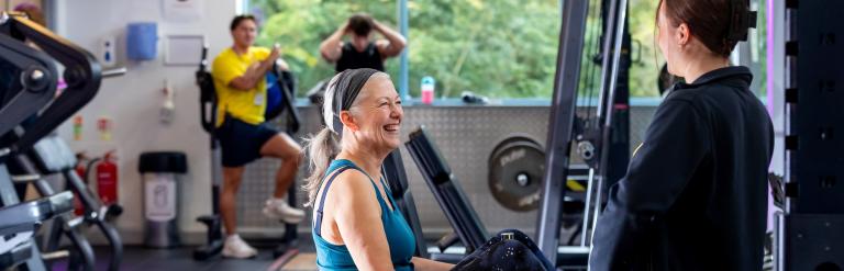 A woman using an exercise machine, smiling at a staff member